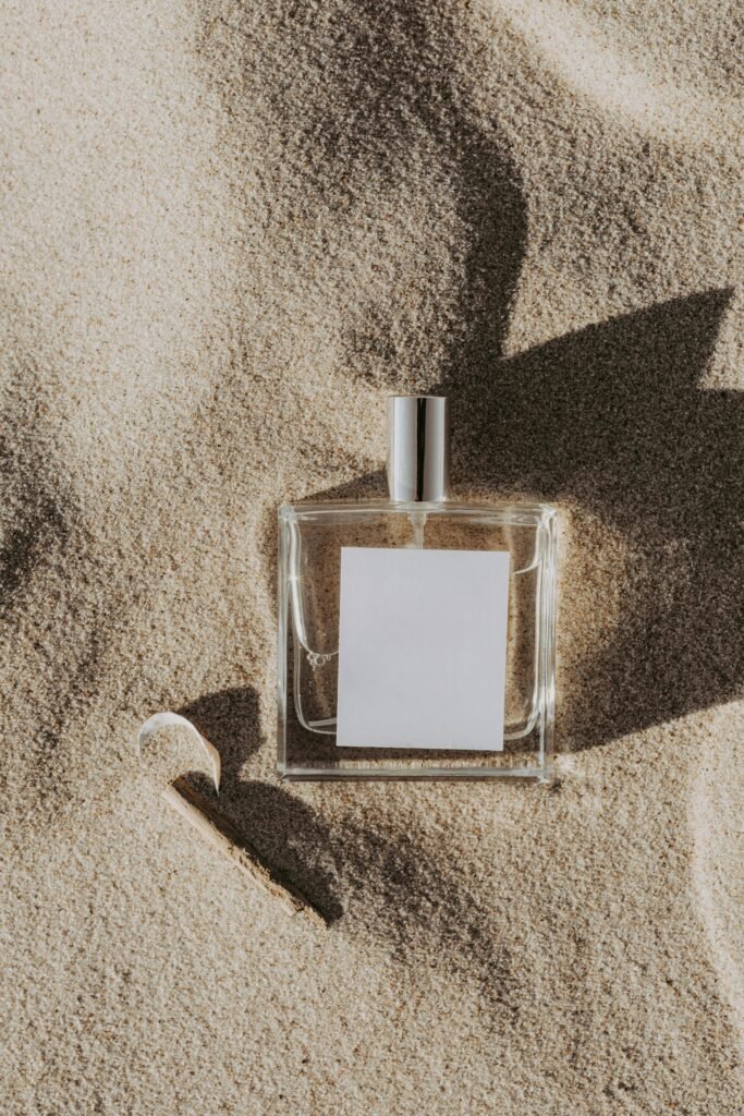 A minimalist perfume bottle with a blank label rests on sand, casting a shadow in daylight.